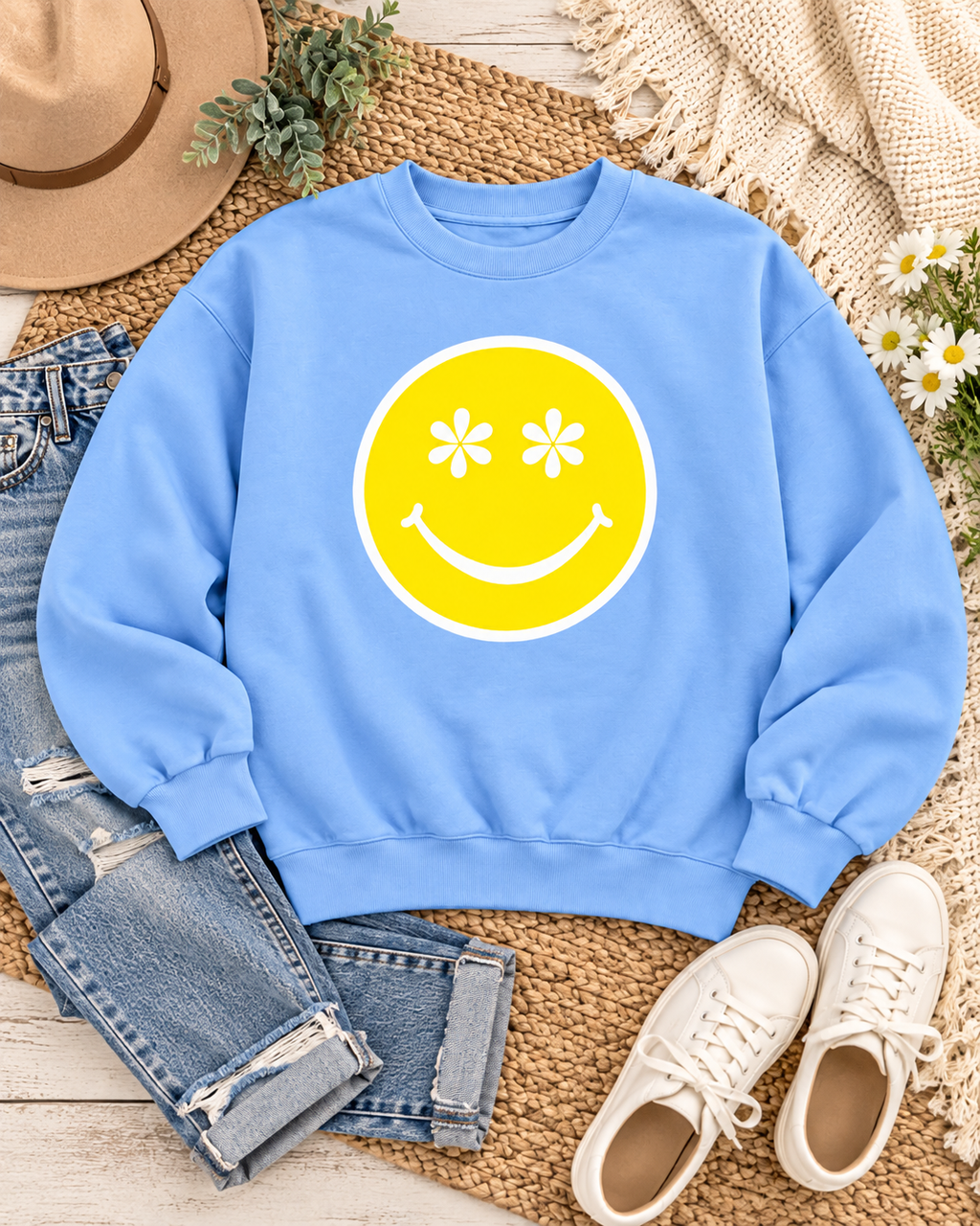 Light blue daisy eyes happy face sweatshirt styled with jeans, cream sneakers, a tan hat, greenery, and daisies
