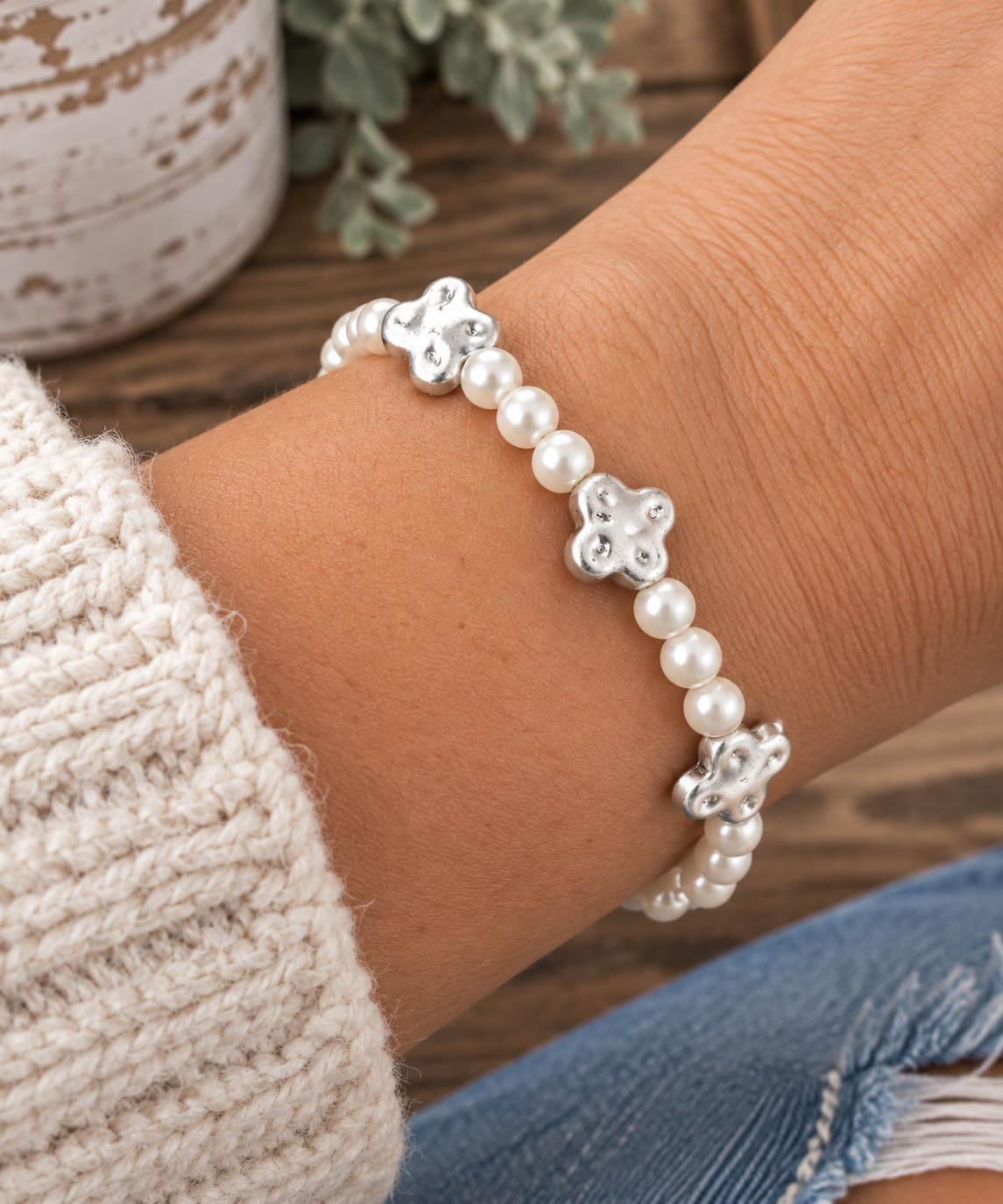 Woman wearing pearl beaded stretch bracelet with silver clover accents on wrist with cream knit sweater, distressed denim, and chippy white pot with eucalyptus backdrop
