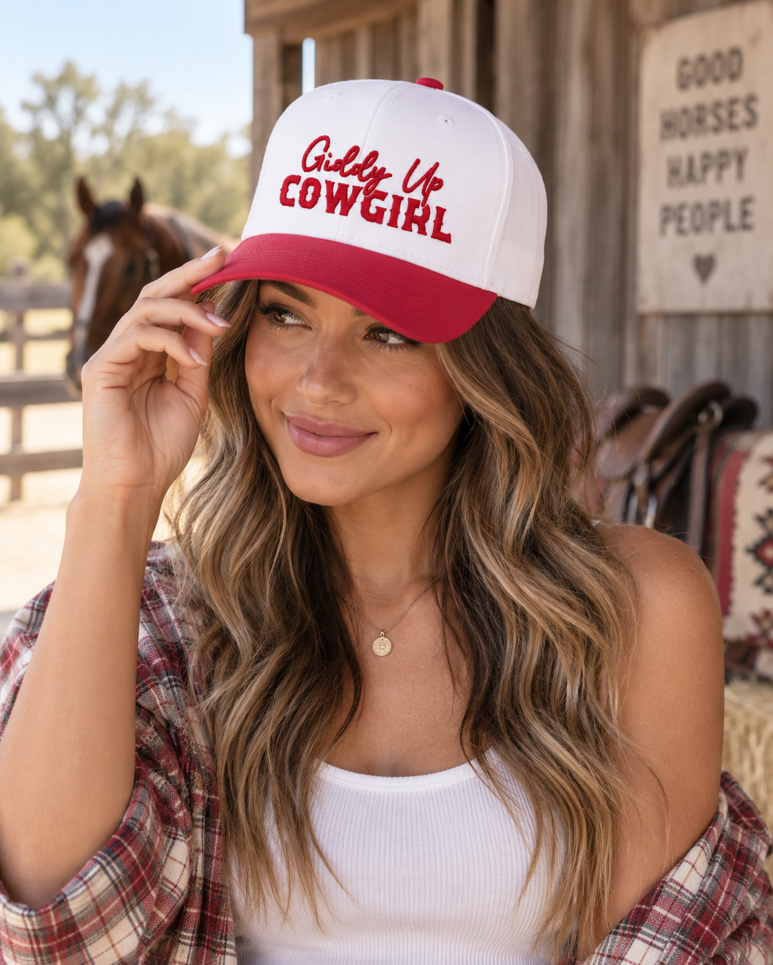 Woman wearing Giddy Up Cowgirl Baseball Cap with white crown and red bill tilted up showing red embroidered Giddy Up Cowgirl lettering with flannel shirt and horse stable backdrop
