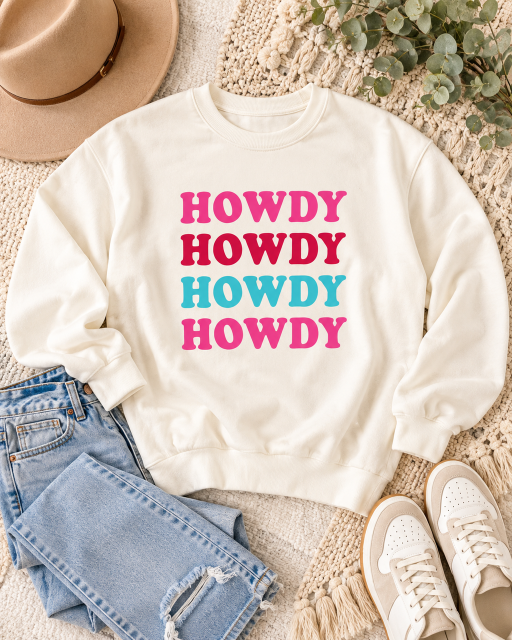 Cream HOWDY sweatshirt styled with blue jeans, white sneakers, a tan hat, and greenery on a textured cream background
