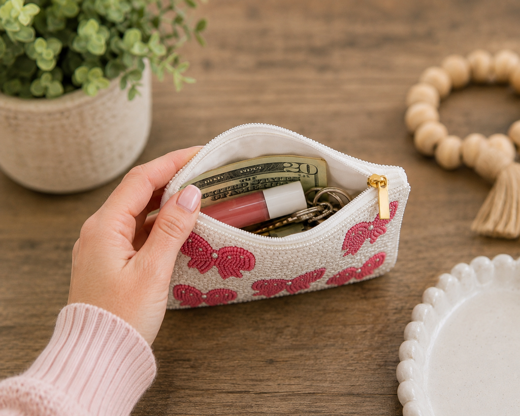 A hand holds an open white zippered pouch with pink embroidered bows, showing it filled with cash, a lip product, and keys, on a wooden tabletop with a plant and bead bracelet in the background.