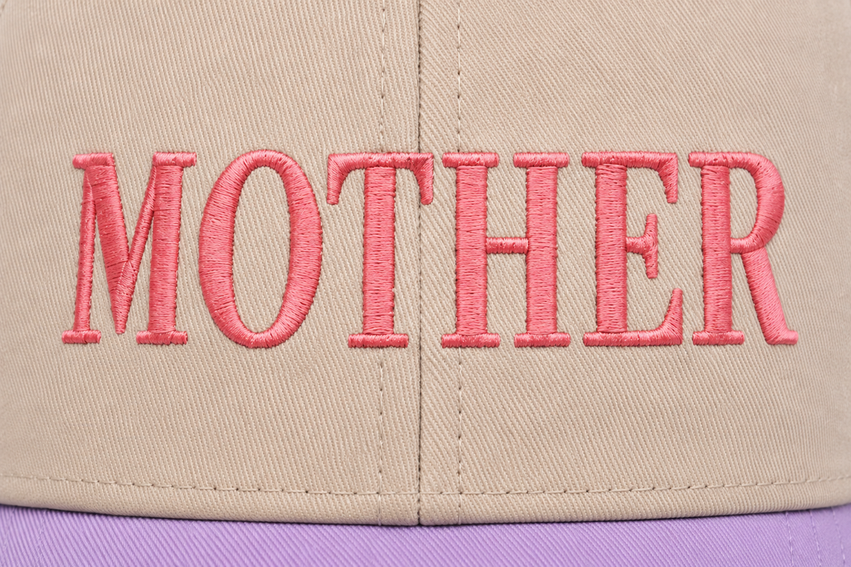 Close-up of bold coral-pink embroidered "MOTHER" lettering in serif style on a khaki baseball cap front panel with lavender brim.

