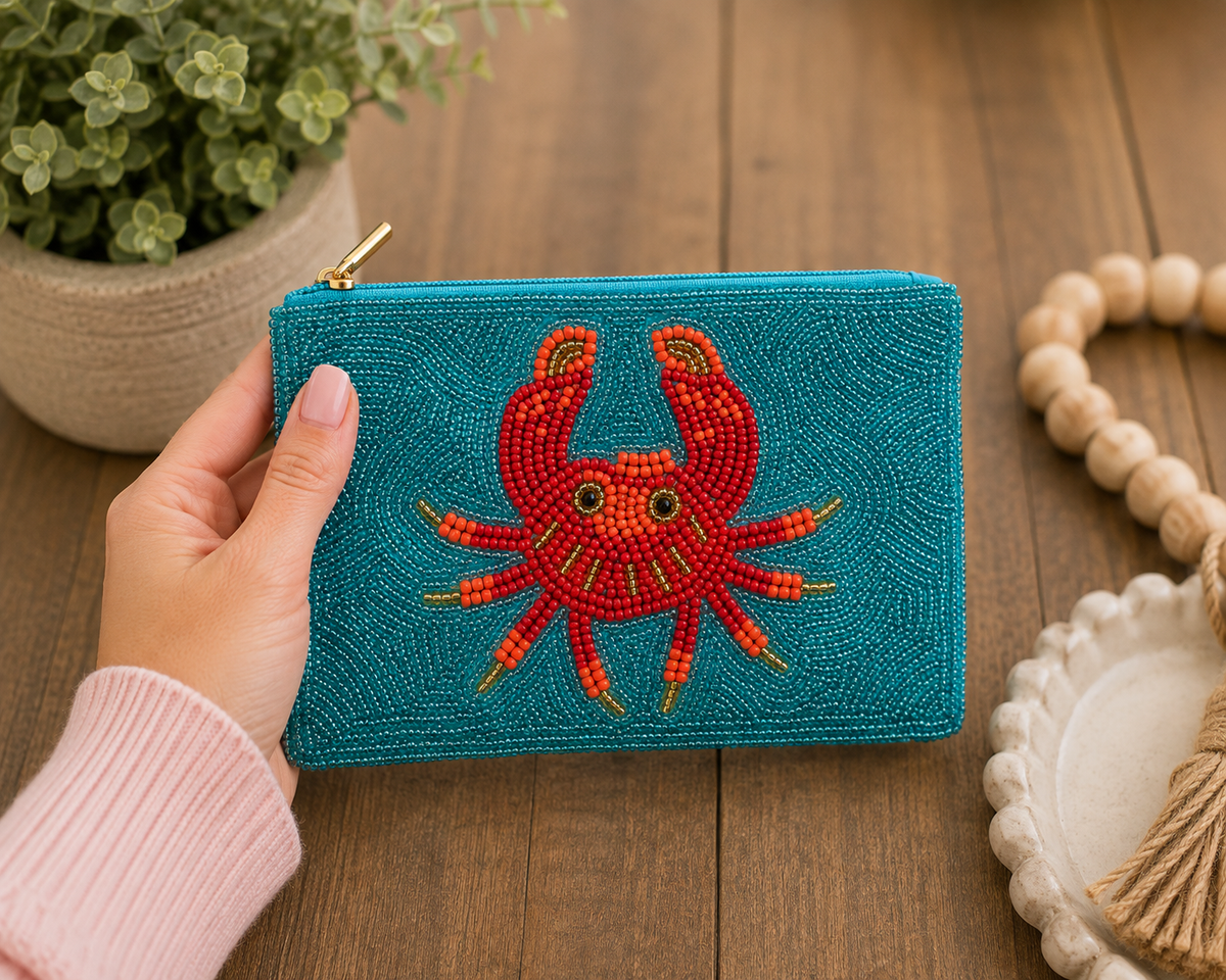 A hand holds a teal beaded zip pouch featuring a large red crab design on the front, set against a rustic wooden table with a potted plant and decorative beads in the background.