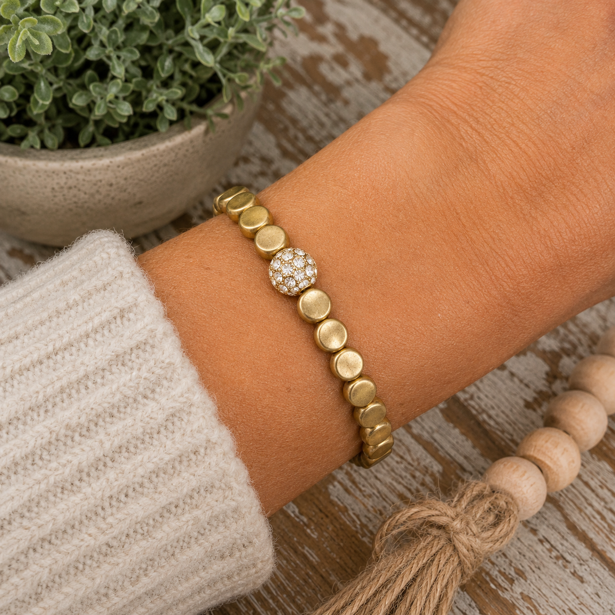 Woman wearing gold coin bead stretch bracelet with crystal accent on wrist with cream knit sweater and wood bead garland
