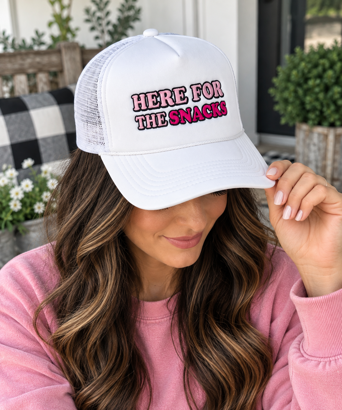 Woman wearing Here for the Snacks Trucker Hat with white front and white mesh back showing pink and hot pink embroidered Here for the Snacks lettering with black outline with pink sweatshirt and outdoor porch backdrop
