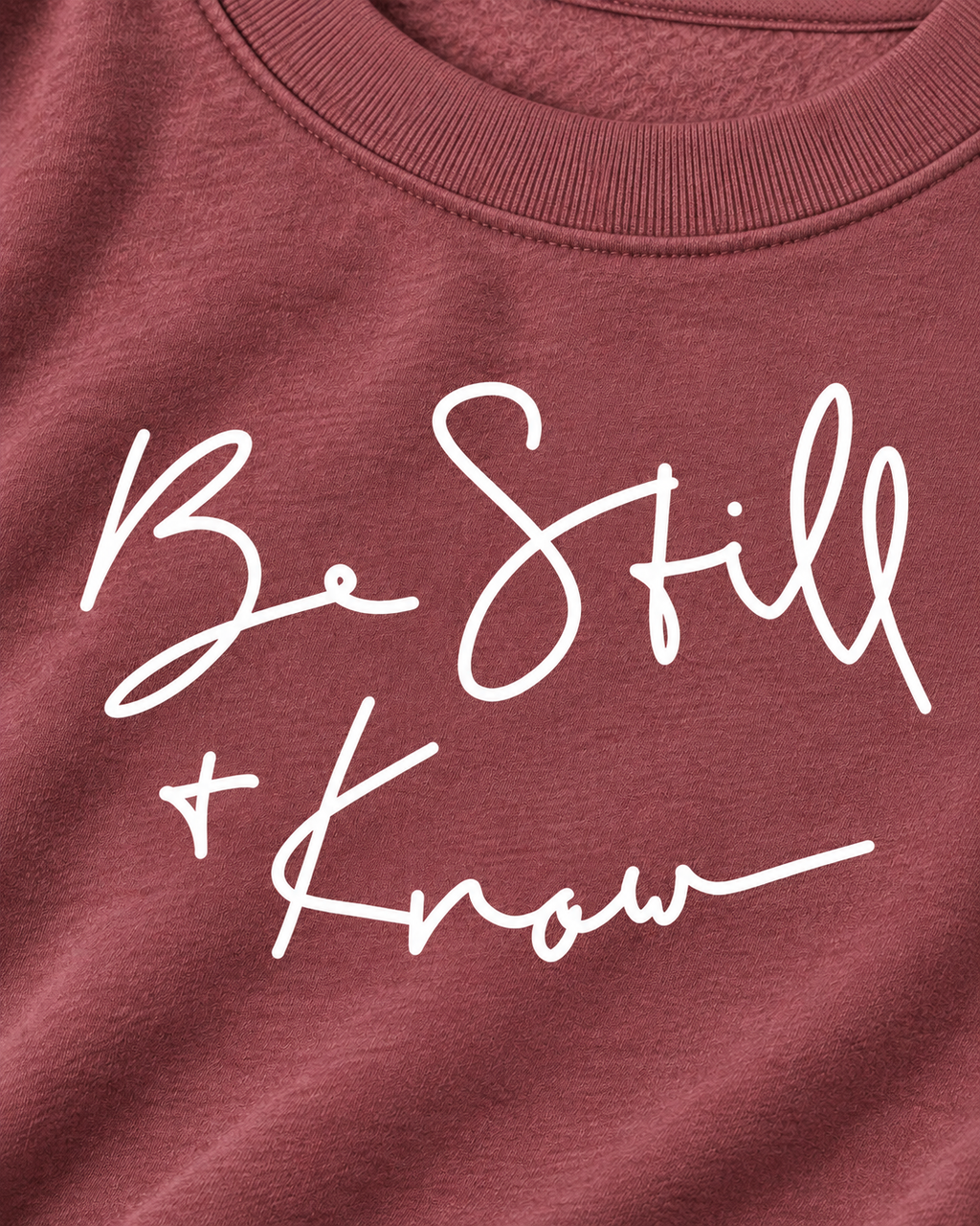 Close-up of white Be Still + Know handwritten-style lettering on the dusty mauve sweatshirt beneath its ribbed neckline
