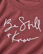 Close-up of white Be Still + Know handwritten-style lettering on the dusty mauve sweatshirt beneath its ribbed neckline