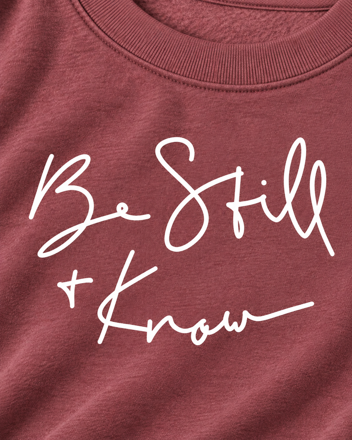 Close-up of white Be Still + Know handwritten-style lettering on the dusty mauve sweatshirt beneath its ribbed neckline