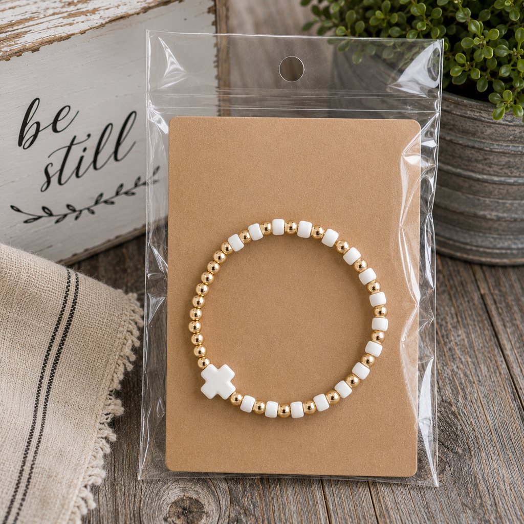 Gold and white cross beaded stretch bracelet on kraft card in clear packaging on rustic wood surface with be still sign and green plant
