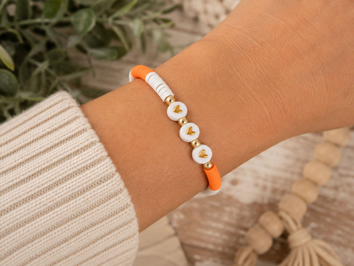 Woman wearing orange and white heart beaded stretch bracelet on wrist with cream knit sweater and greenery background
