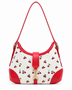 Cherry Pattern Hamptons Hobo Bag front view showing cream canvas body with allover red cherry print, red faux leather curved top trim and corner accents, center red strap tab with gold horsebit snaffle clasp, and single red faux leather shoulder strap with gold D-ring attachments on white background

