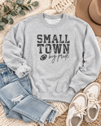 Small Town Big Pride football sweatshirt styled with jeans, sneakers, and a tan hat on a cream blanket
