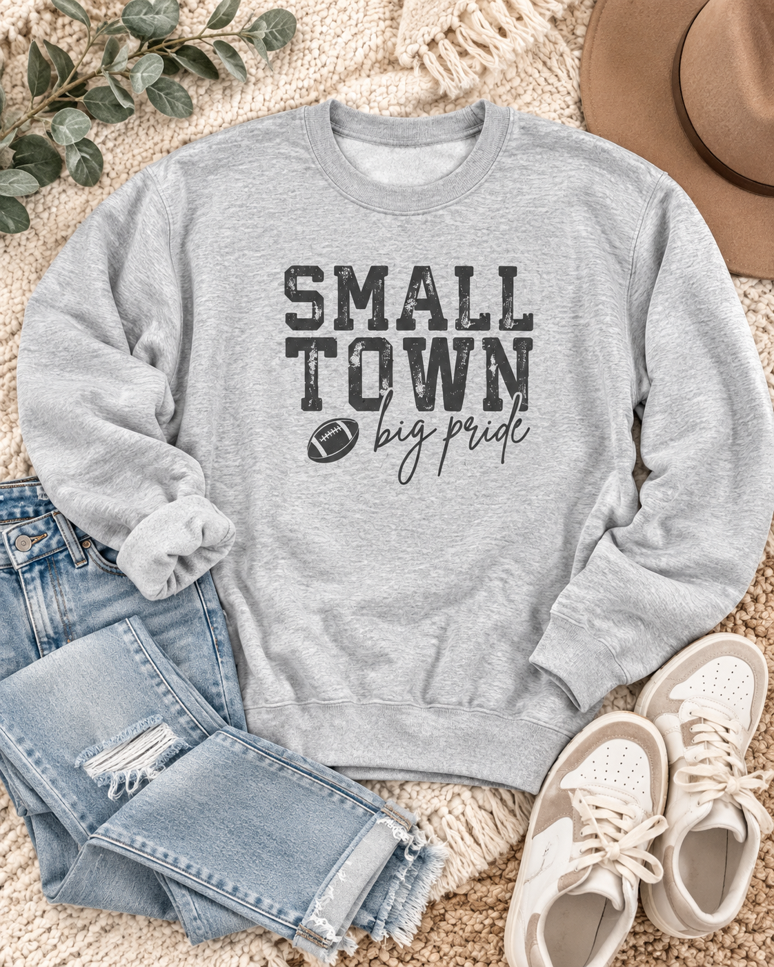 Small Town Big Pride football sweatshirt styled with jeans, sneakers, and a tan hat on a cream blanket
