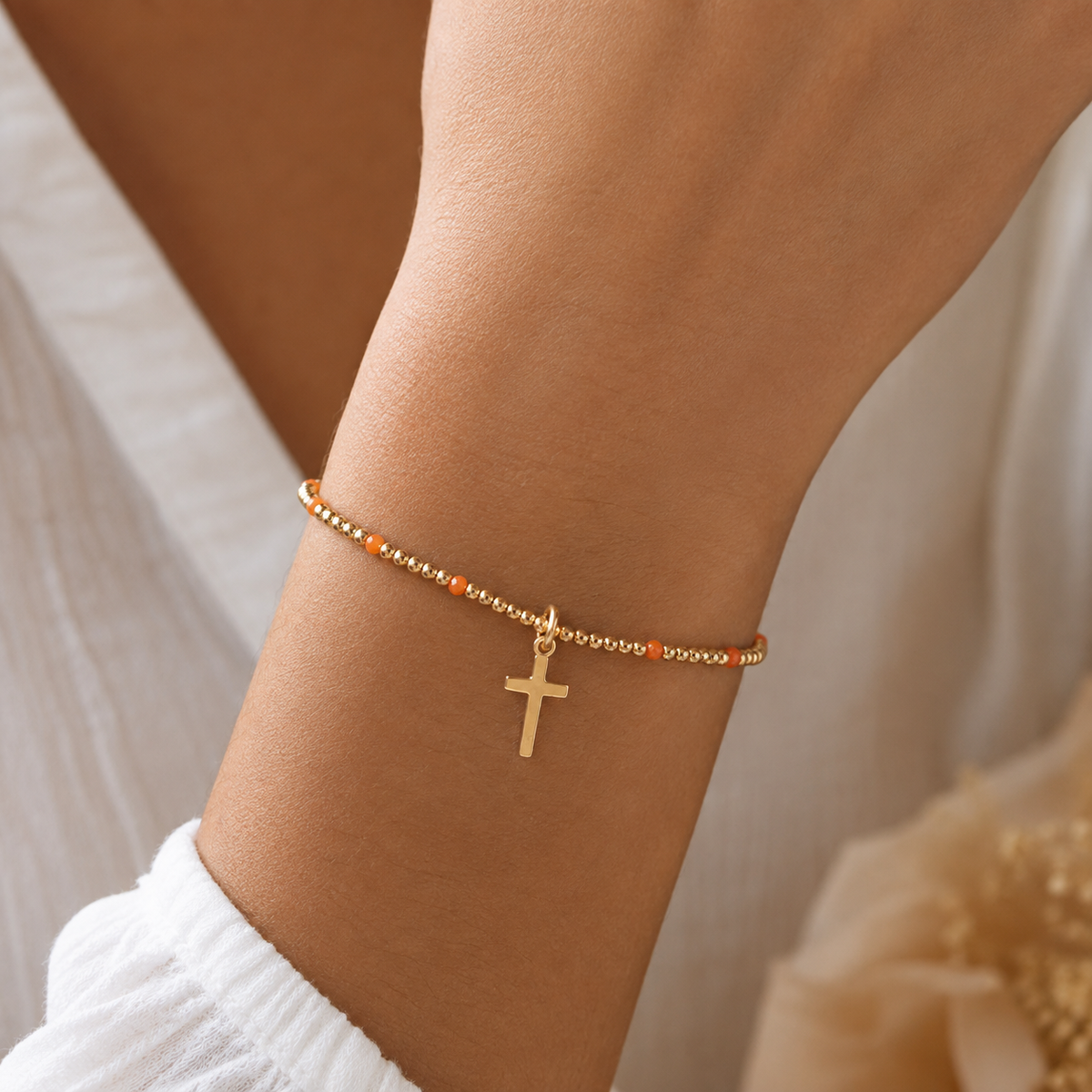 Woman wearing gold beaded cross bracelet with orange accents and gold cross charm on wrist with white knit sleeve
