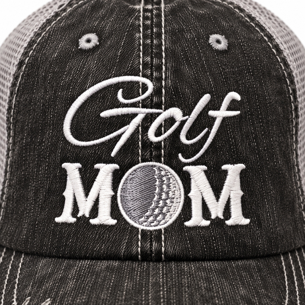 Close-up of white embroidered Golf script and Mom block lettering with gray embroidered golf ball detail replacing the letter O on distressed black trucker hat front panel
