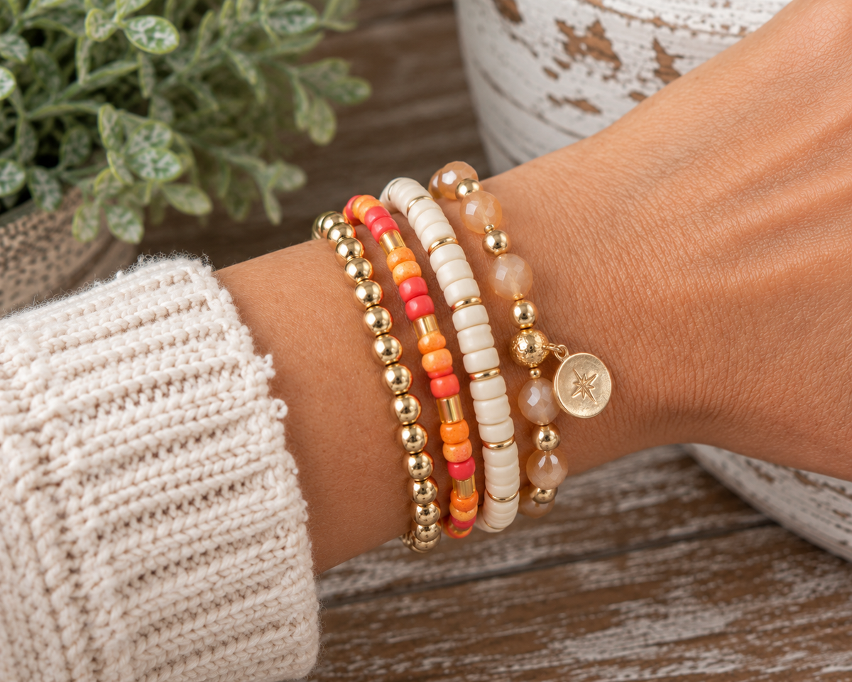 Coral and orange seed bead bracelet stacked with white seed bead bracelet, gold ball bracelet, and stone charm bracelet on wrist with cream knit sleeve and rustic wood backdrop
