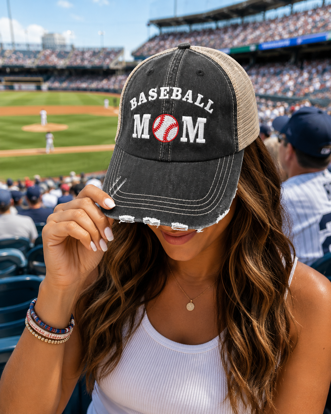 A woman at a baseball game wearing a distressed black trucker hat with Baseball Mom in white text and a baseball graphic on the front, with a blurred stadium background.
