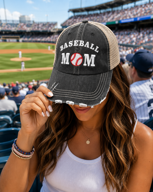 A woman at a baseball game wearing a distressed black trucker hat with Baseball Mom in white text and a baseball graphic on the front, with a blurred stadium background.