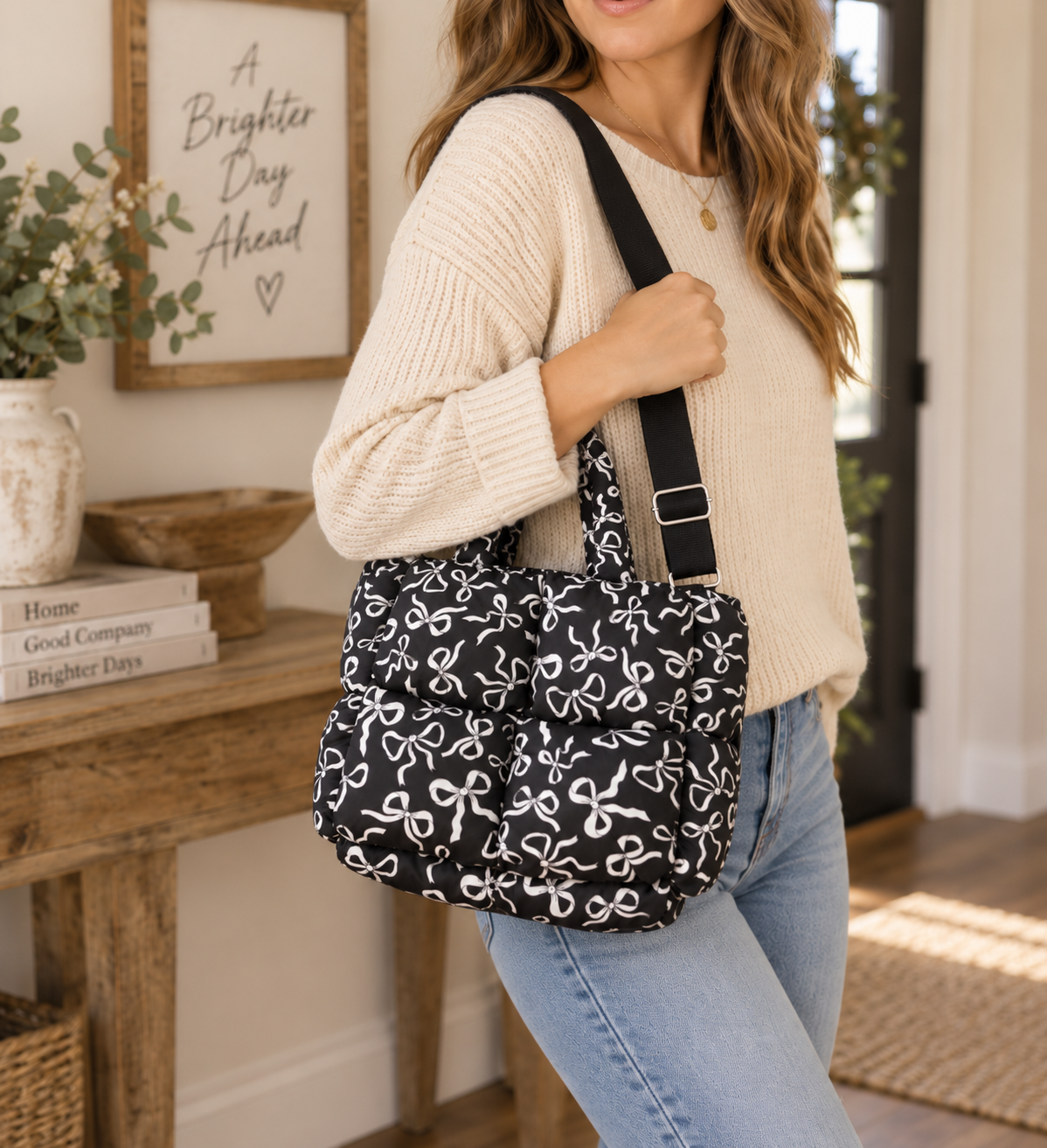 Woman carrying Black Bow Print Puffer Tote on shoulder showing black puffer body with all-over white bow print, adjustable black crossbody strap, and dual top handles with cream sweater in bright farmhouse entryway
