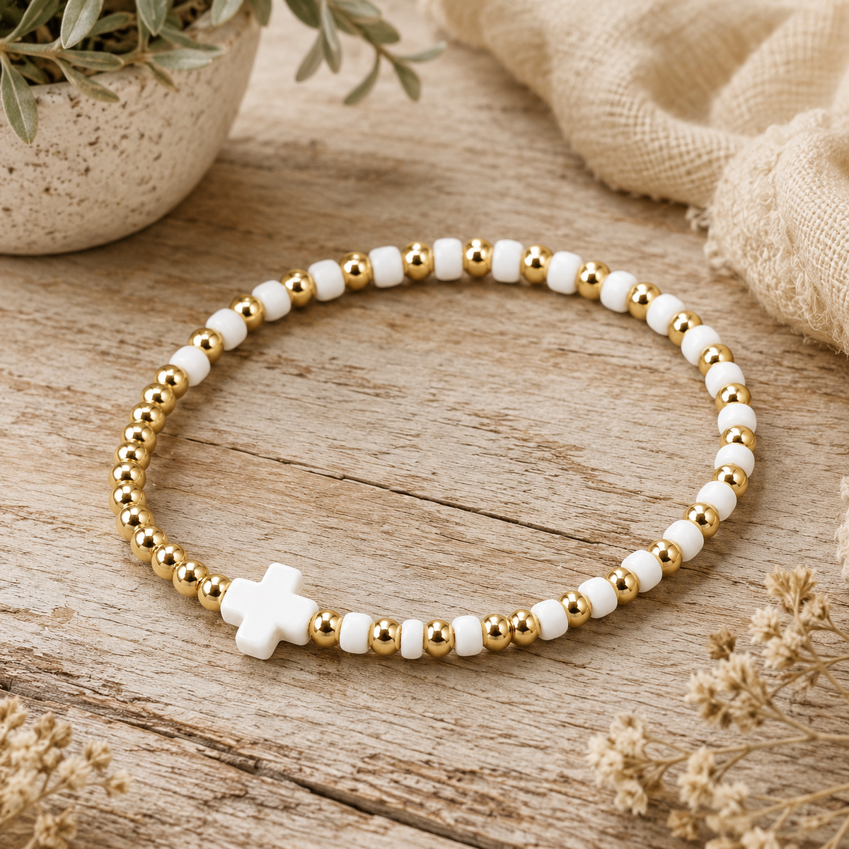 Gold and white cross beaded stretch bracelet laid flat on rustic wood surface with dried flowers, ceramic pot, and linen fabric backdrop
