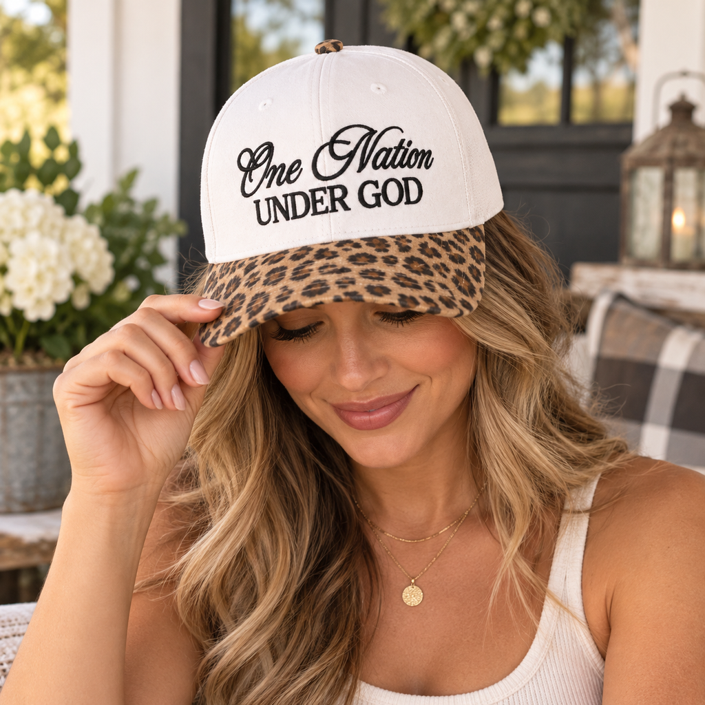 Blonde woman tilting brim of One Nation Under God Baseball Cap in Leopard colorway showing white structured crown with black embroidered script and tan and black leopard print bill in boutique Western shop setting with chalkboard sign and brown felt hat in background
