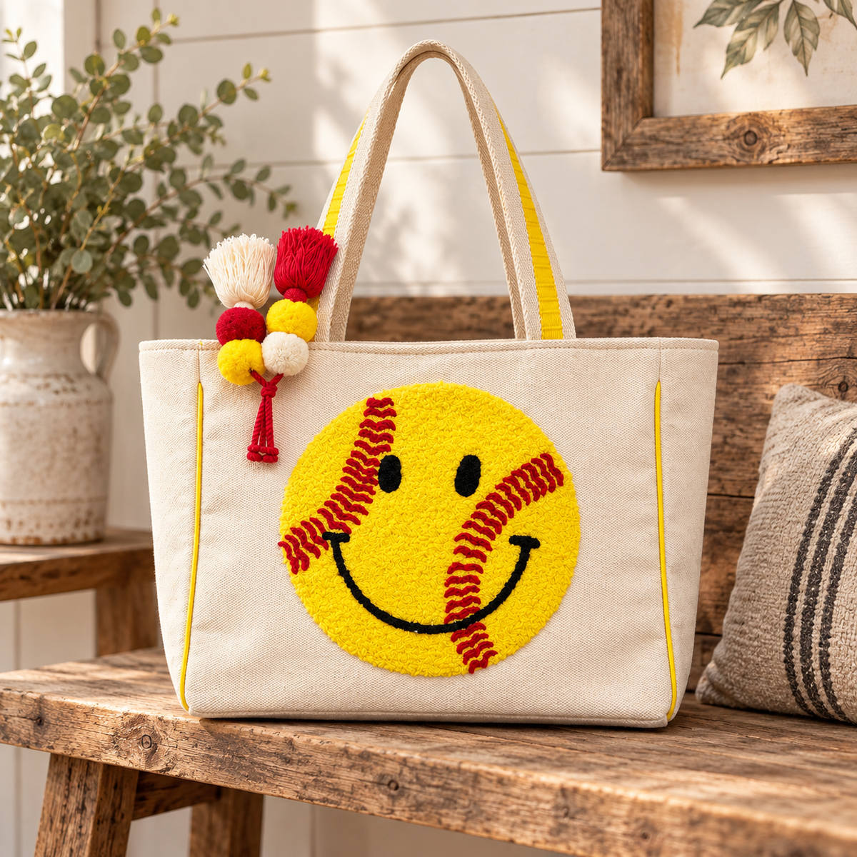 Softball Smiley Tote Bag displayed on rustic wooden bench showing natural canvas body with large yellow textured softball smiley face, red stitching, yellow trim, and colorful pom-pom tassel with farmhouse backdrop
