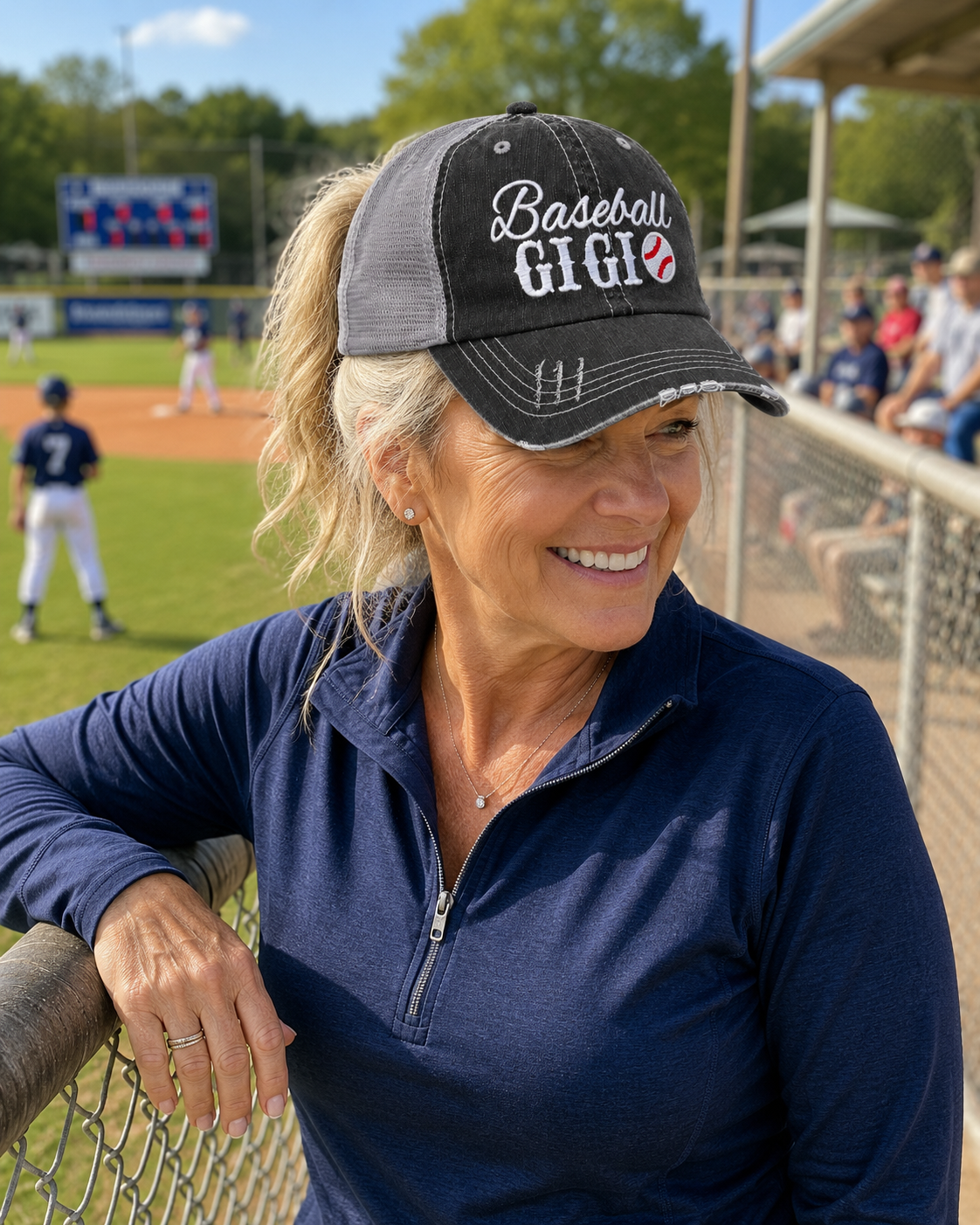 A smiling woman at a baseball game wearing a black and gray trucker hat with "Baseball GIGI" embroidered on the front, leaning on a fence in front of a baseball field.
