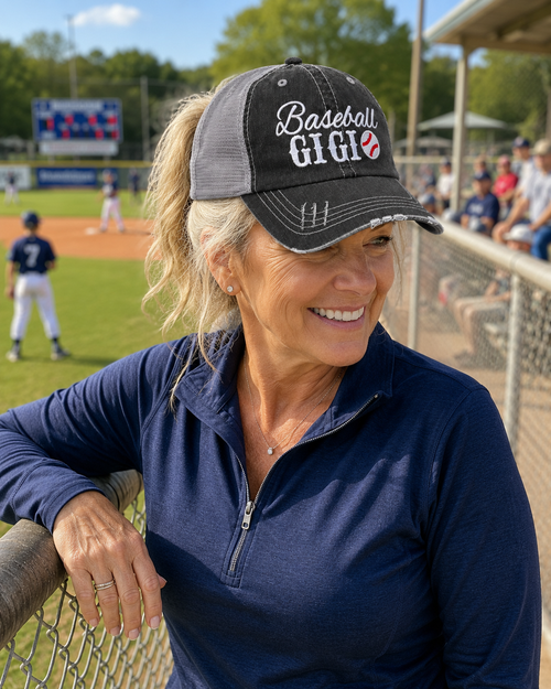 A smiling woman at a baseball game wearing a black and gray trucker hat with "Baseball GIGI" embroidered on the front, leaning on a fence in front of a baseball field.

