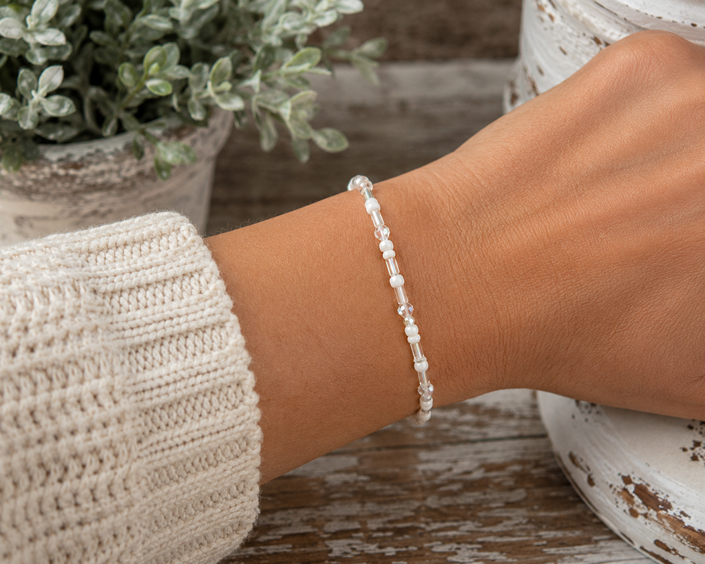 Woman wearing white and iridescent seed bead stretch bracelet on wrist with cream knit sweater and rustic chippy white pedestal backdrop
