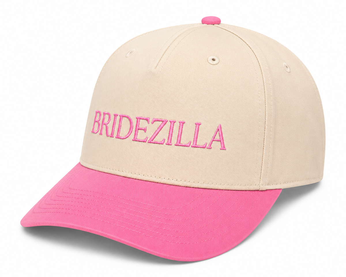 Close-up of bold pink embroidered "BRIDEZILLA" lettering on a cream baseball cap front panel with hot pink brim.
