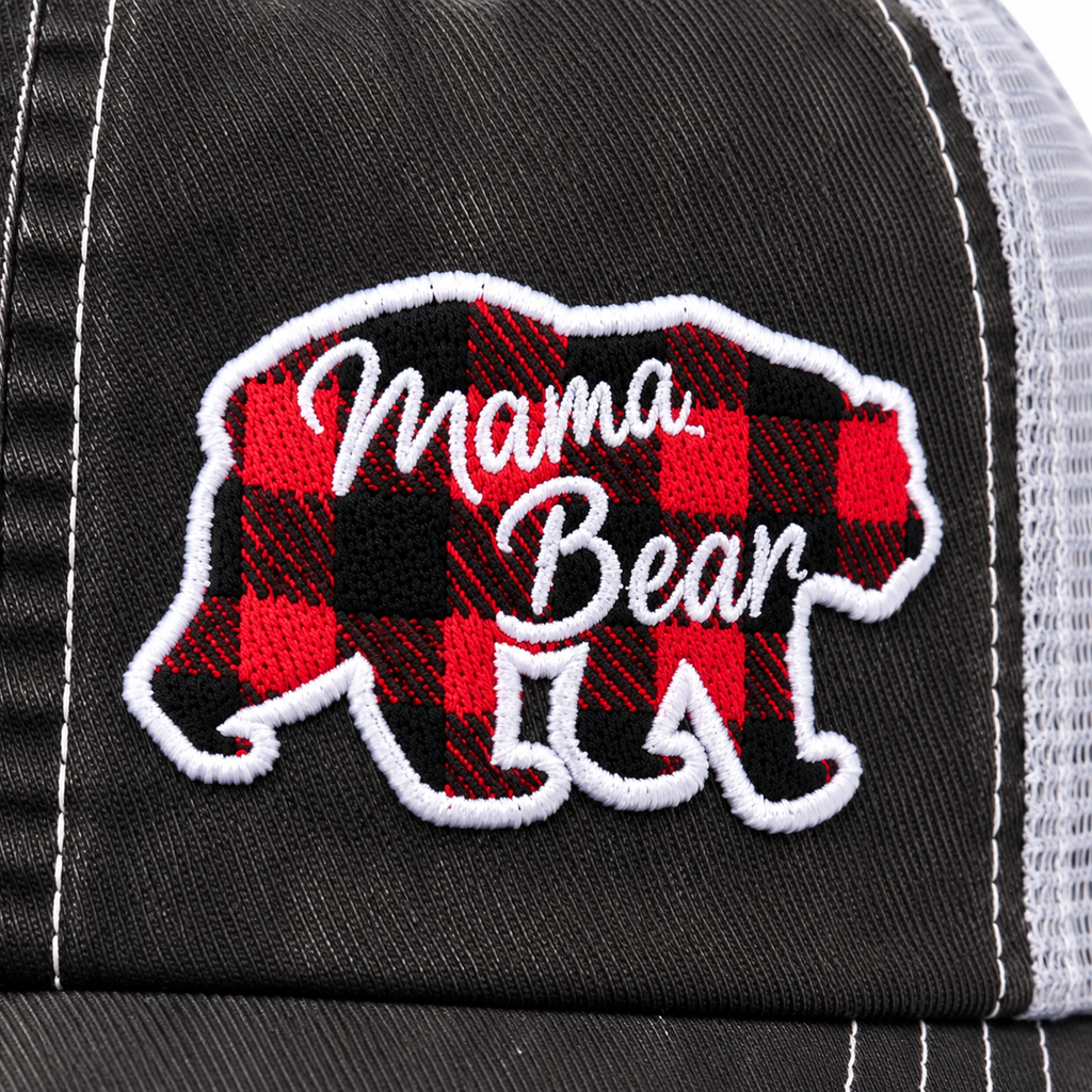 Close-up of red and black buffalo plaid embroidered bear patch with white Mama Bear script lettering and white border outline on distressed black trucker hat front panel
