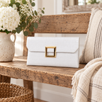 White Croc Envelope Clutch on rustic reclaimed wood bench showing white croc-embossed vegan leather and open gold square hardware beside white hydrangeas and baby's breath in cream speckled ceramic pitcher, cream and grey stripe linen throw, and wicker basket with gilt mirror in background
