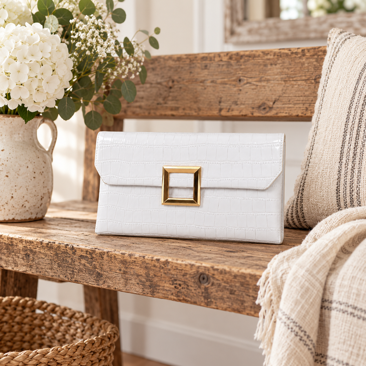 White Croc Envelope Clutch on rustic reclaimed wood bench showing white croc-embossed vegan leather and open gold square hardware beside white hydrangeas and baby's breath in cream speckled ceramic pitcher, cream and grey stripe linen throw, and wicker basket with gilt mirror in background
