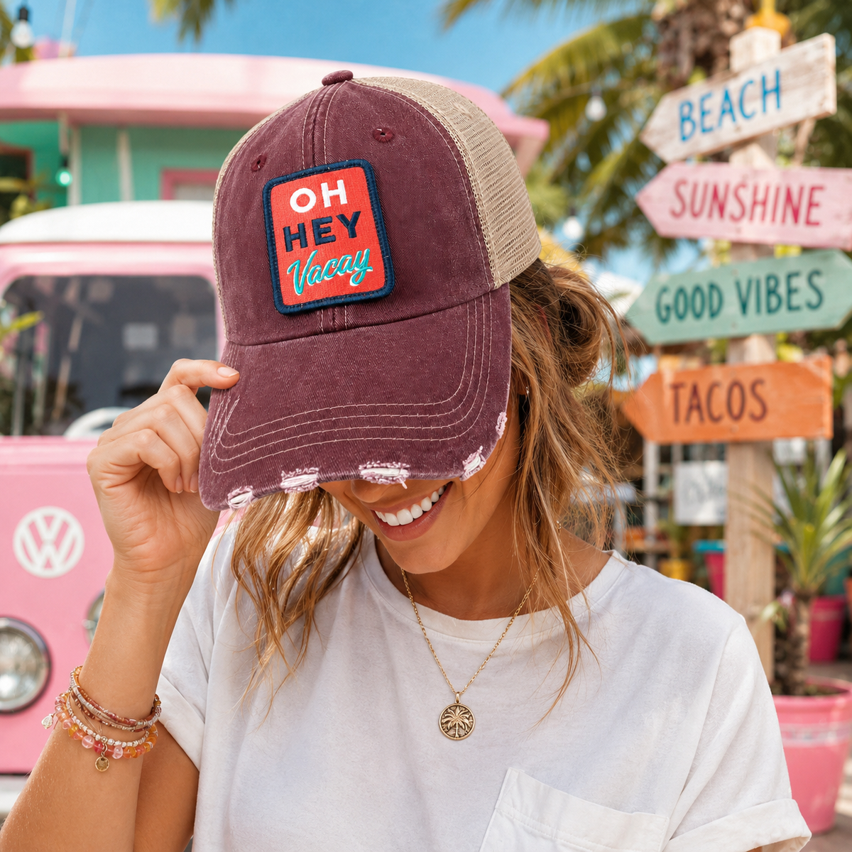 A woman wearing a maroon and beige trucker-style baseball cap with a colorful 'OHHEY Vacay' patch on the front, posing outdoors in front of a pink VW van and a colorful signpost.