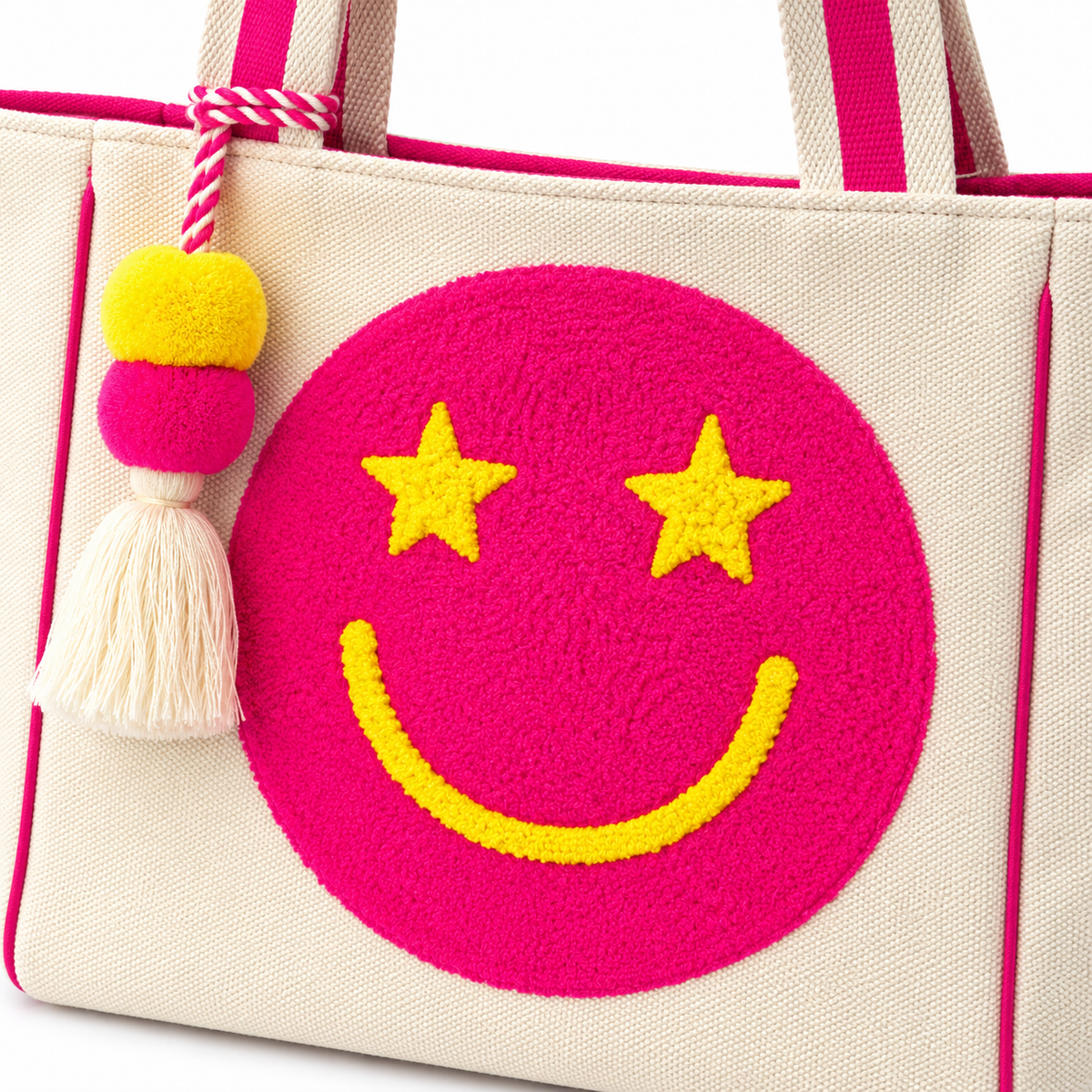 Close-up of Pink Star Smiley Canvas Tote Bag showing oversized hot pink chenille smiley face patch with yellow star eyes and yellow smile, and pom-pom tassel charm with yellow pom, hot pink pom, and cream fringe tassel on red and white twisted cord
