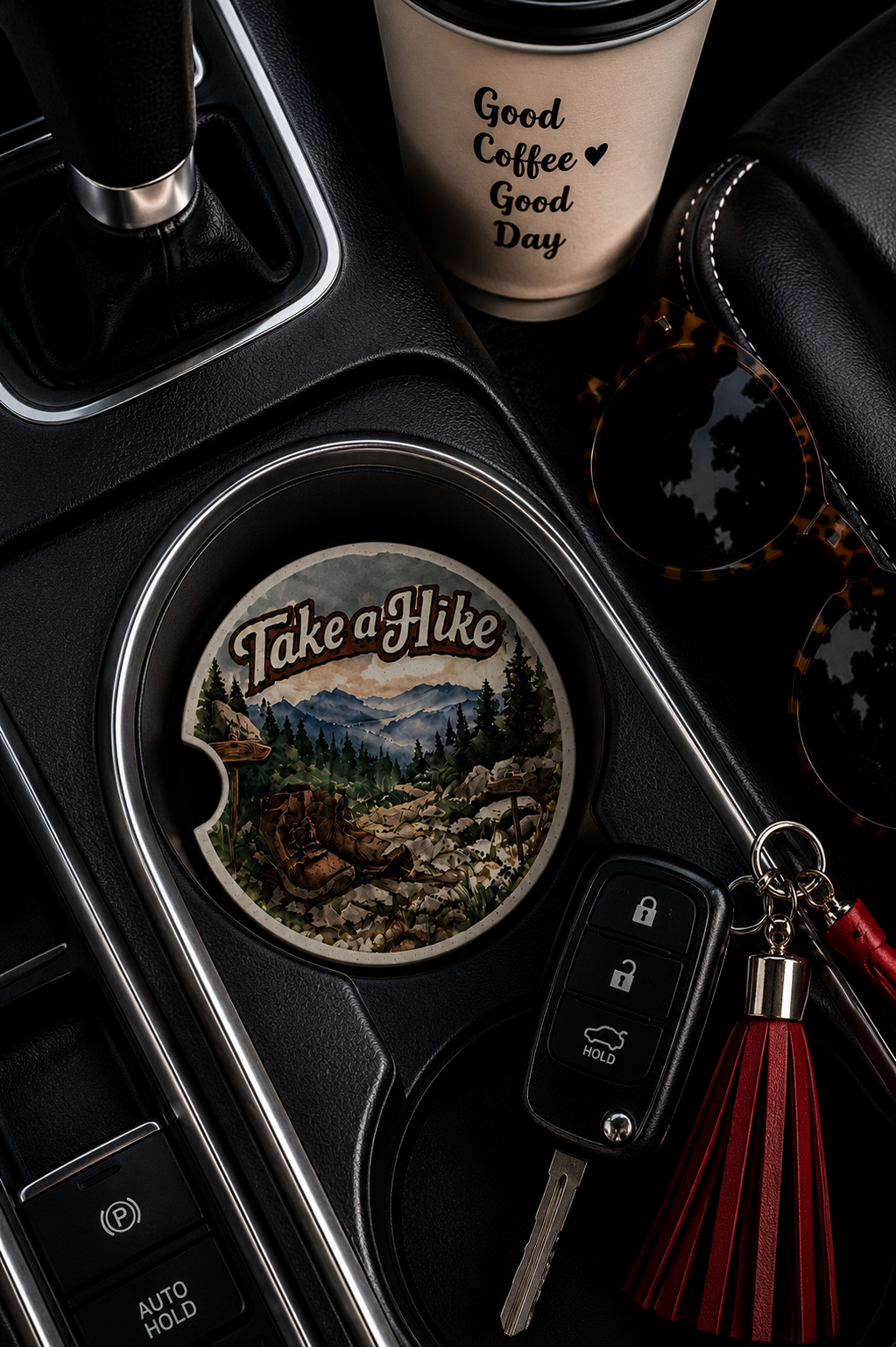 Take a Hike ceramic car coaster in cup holder — watercolor mountain trail with hiking boots, pine trees, and misty blue mountains, shown in car console with coffee cup and sunglasses
