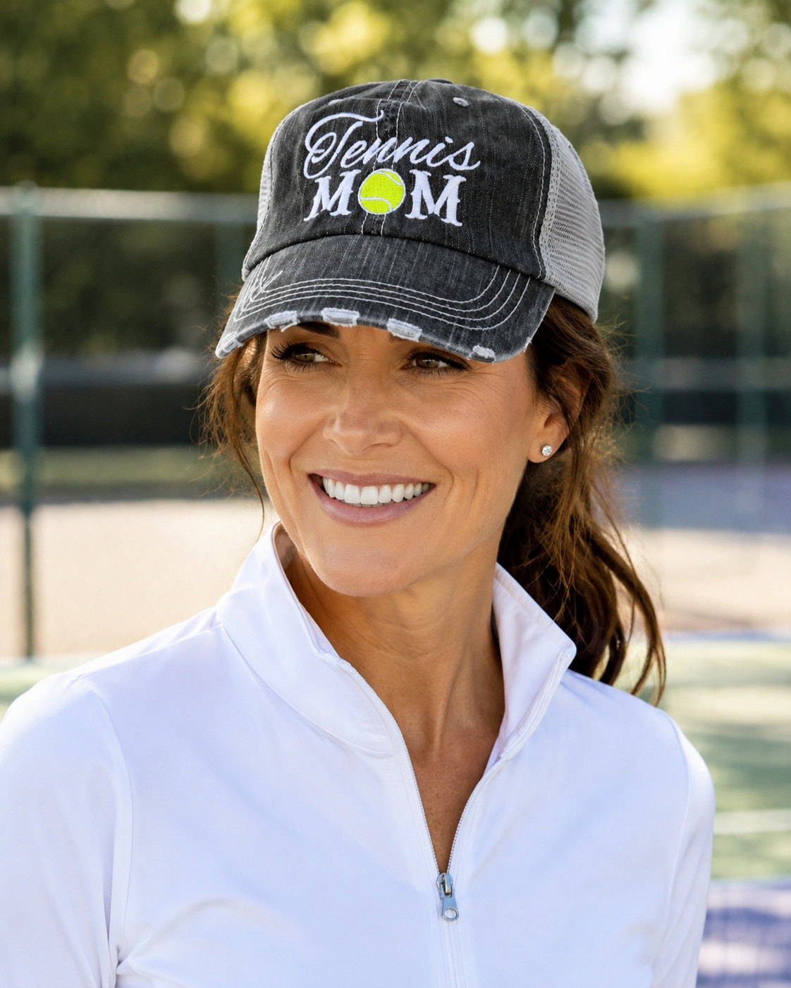 Woman wearing the Tennis Mom Trucker Hat on a tennis court, smiling, with the embroidered design and distressed charcoal brim visible
