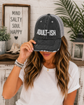 A woman wearing a distressed trucker-style baseball cap with 'ADULT-ISH' printed in white on a dark gray washed front panel and mesh back. She is holding the brim of the hat, posing in a styled indoor setting with a rustic sign and plants in the background.
