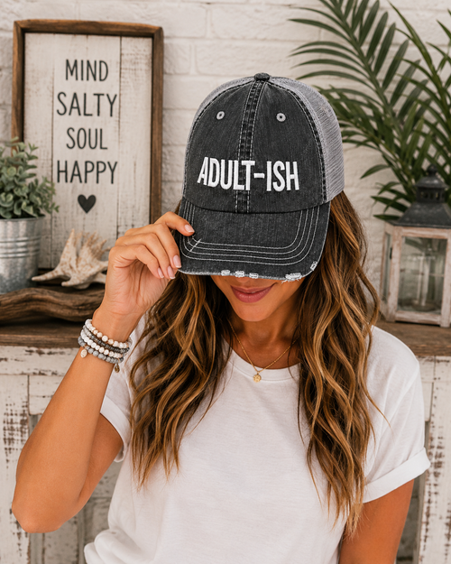 A woman wearing a distressed trucker-style baseball cap with 'ADULT-ISH' printed in white on a dark gray washed front panel and mesh back. She is holding the brim of the hat, posing in a styled indoor setting with a rustic sign and plants in the background.