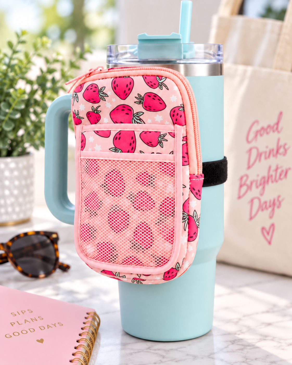 Strawberry Tumbler Pouch attached to teal tumbler handle with black elastic strap showing light pink strawberry and star print, pink top zipper, and front mesh pocket beside pink journal, sunglasses, and Good Drinks Brighter Days tote on marble surface
