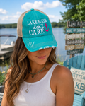 A woman wearing a turquoise and beige trucker-style baseball cap with the text 'LAKE HAIR don't CARE' and a small pink anchor graphic, photographed outdoors near water and a blue chair.