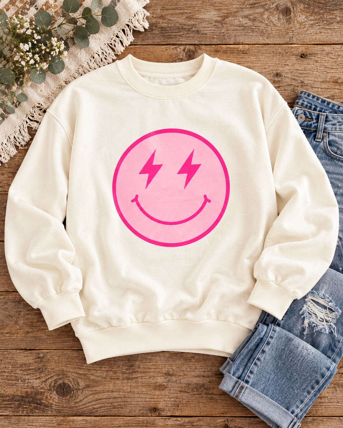 Cream lightning-bolt smiley sweatshirt styled with distressed jeans, flowers, greenery, and a fringed throw on rustic wood
