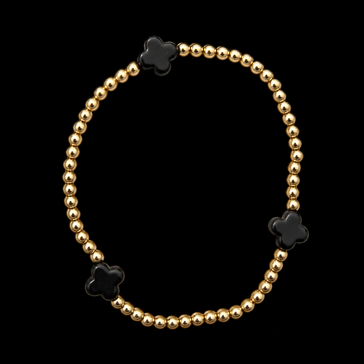 Gold beaded stretch bracelet with three black clover accents displayed flat in a circle on black background

