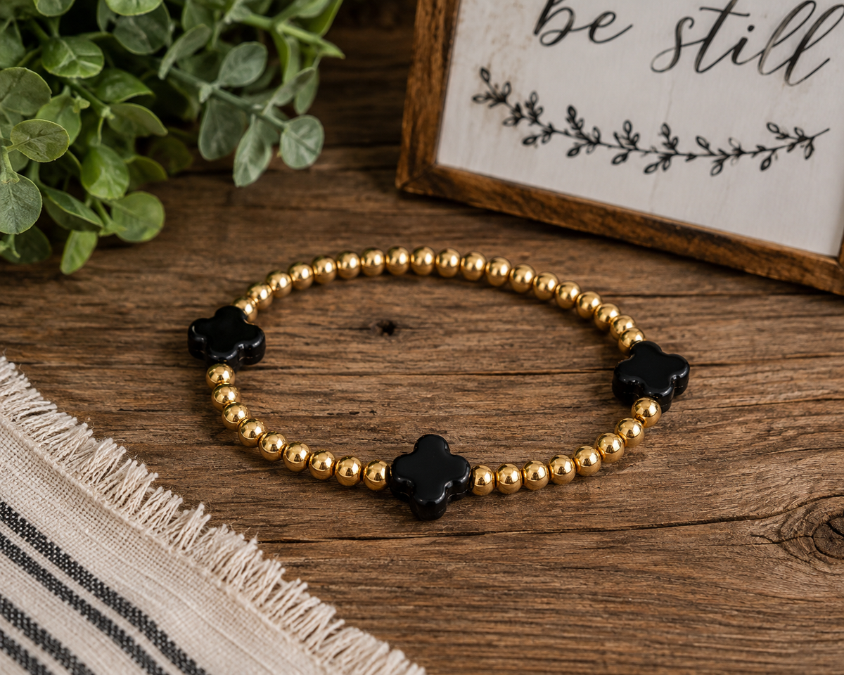 Gold and black clover beaded stretch bracelet laid flat on rustic wood surface with potted greenery, striped linen, and be still sign backdrop
