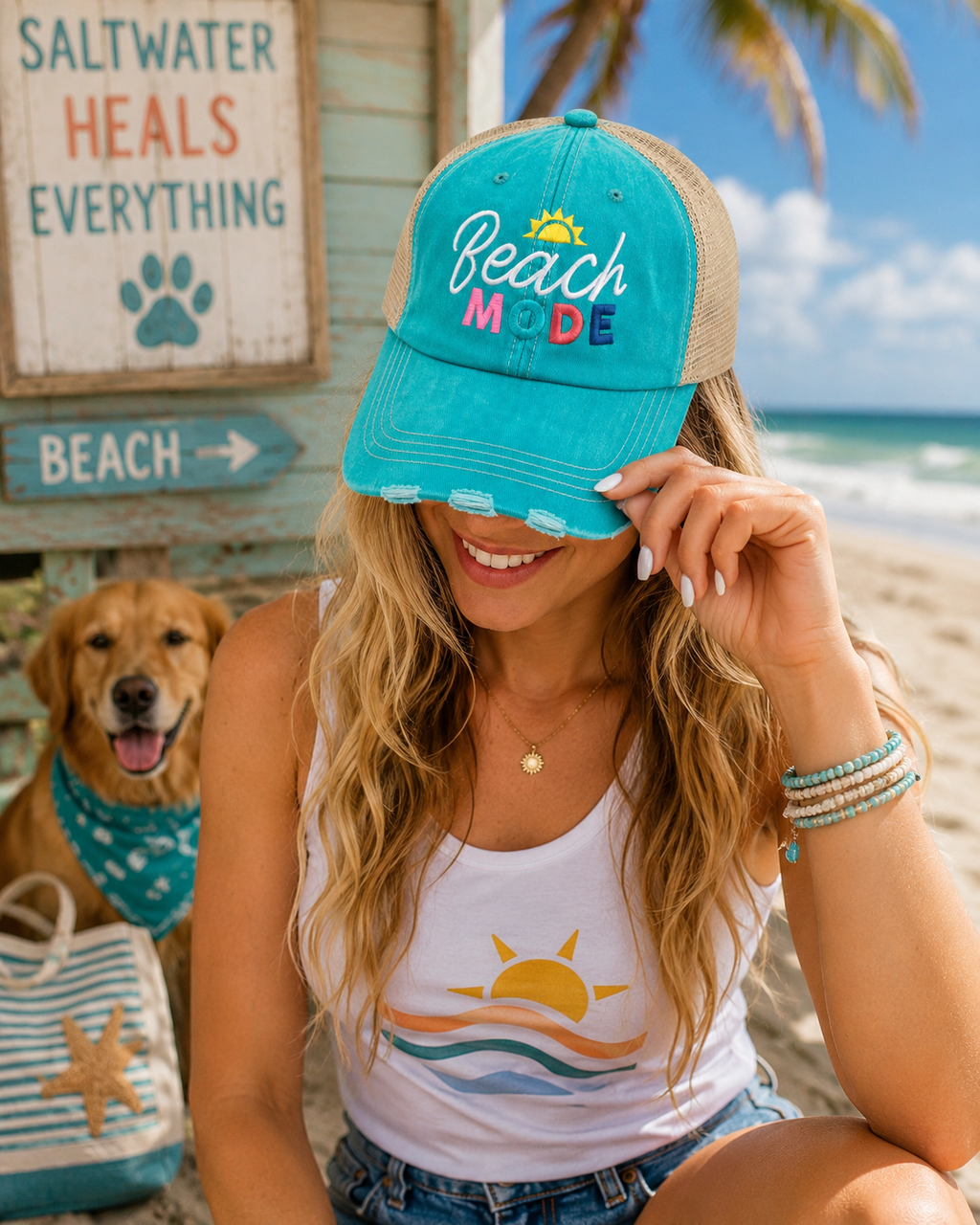 A woman at the beach wearing a turquoise and tan trucker hat with 'Beach Mode' embroidered on the front, alongside a golden retriever and a beach-themed tote bag.