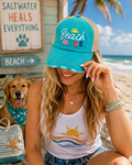 A woman at the beach wearing a turquoise and tan trucker hat with 'Beach Mode' embroidered on the front, alongside a golden retriever and a beach-themed tote bag.
