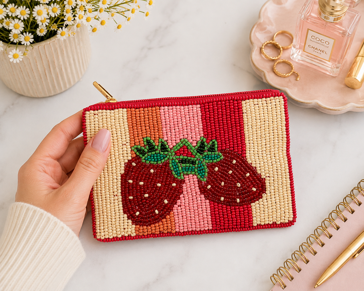 A hand holds a small rectangular beaded pouch with a zipper, decorated with two red strawberries on a striped background of red, orange, pink, and cream. The pouch is photographed on a light marble surface with decorative props including a Chanel perfume bottle, rings, a notebook, and a vase of daisies.