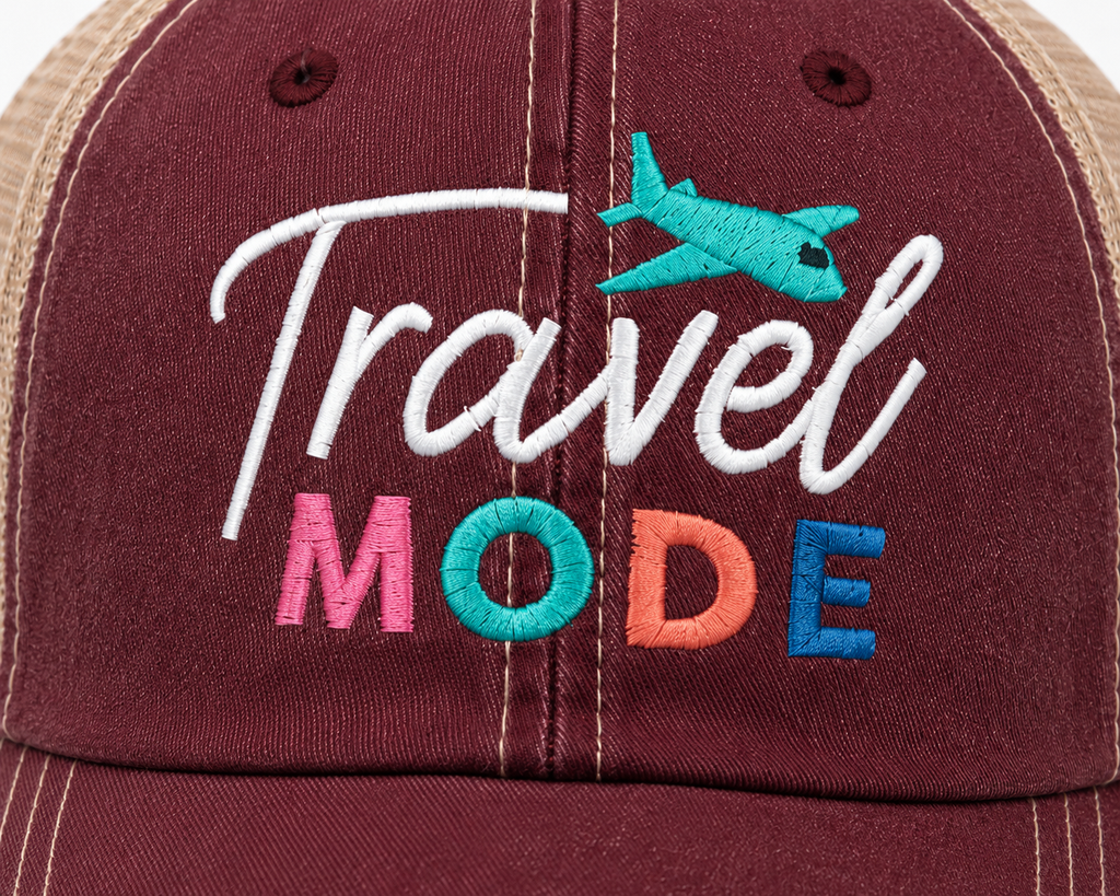 Close-up of Travel Mode trucker hat showing colorful embroidered lettering and turquoise airplane accent on distressed burgundy denim.