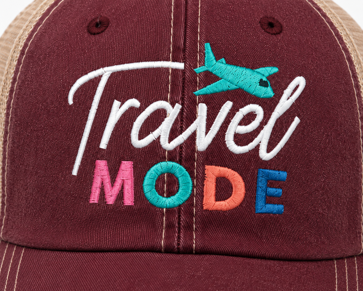 Close-up of Travel Mode trucker hat showing colorful embroidered lettering and turquoise airplane accent on distressed burgundy denim.