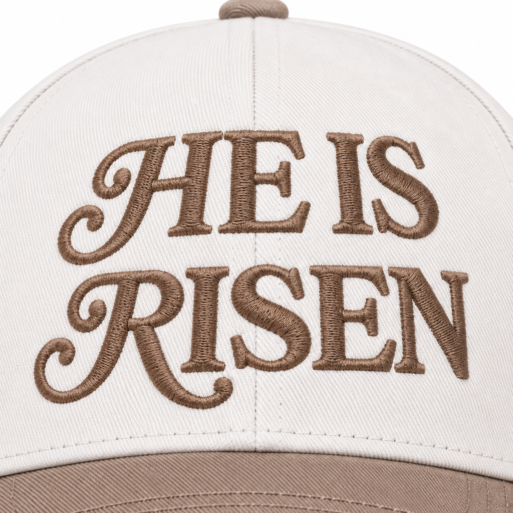 Close-up of He Is Risen Baseball Cap in Ash Mocha showing mocha raised decorative serif script He Is Risen embroidery on cream structured cotton crown with mocha bill edge visible at bottom on white background
